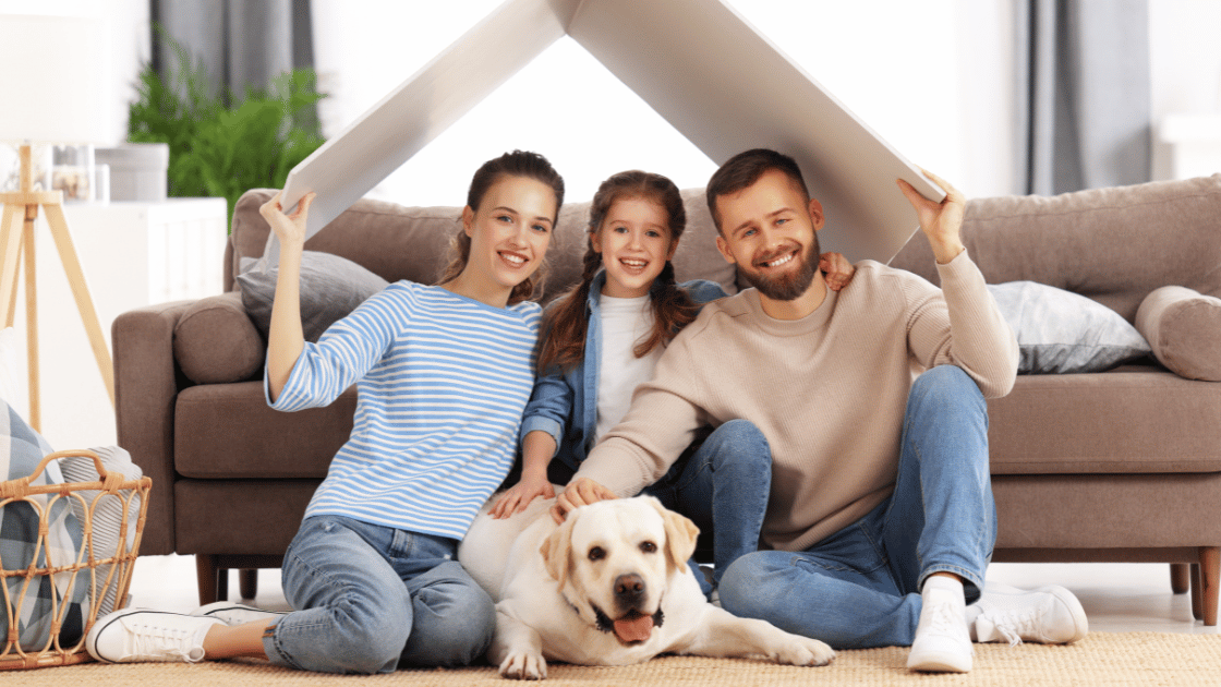 Smiling family with a young daughter and their dog sitting on the floor under a cardboard roof, symbolising their new home.